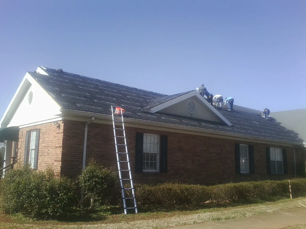 Crew tearing shingles off a brick ranch roof with a ladder set — Metal Roof Repair work by our crew in Sherburne