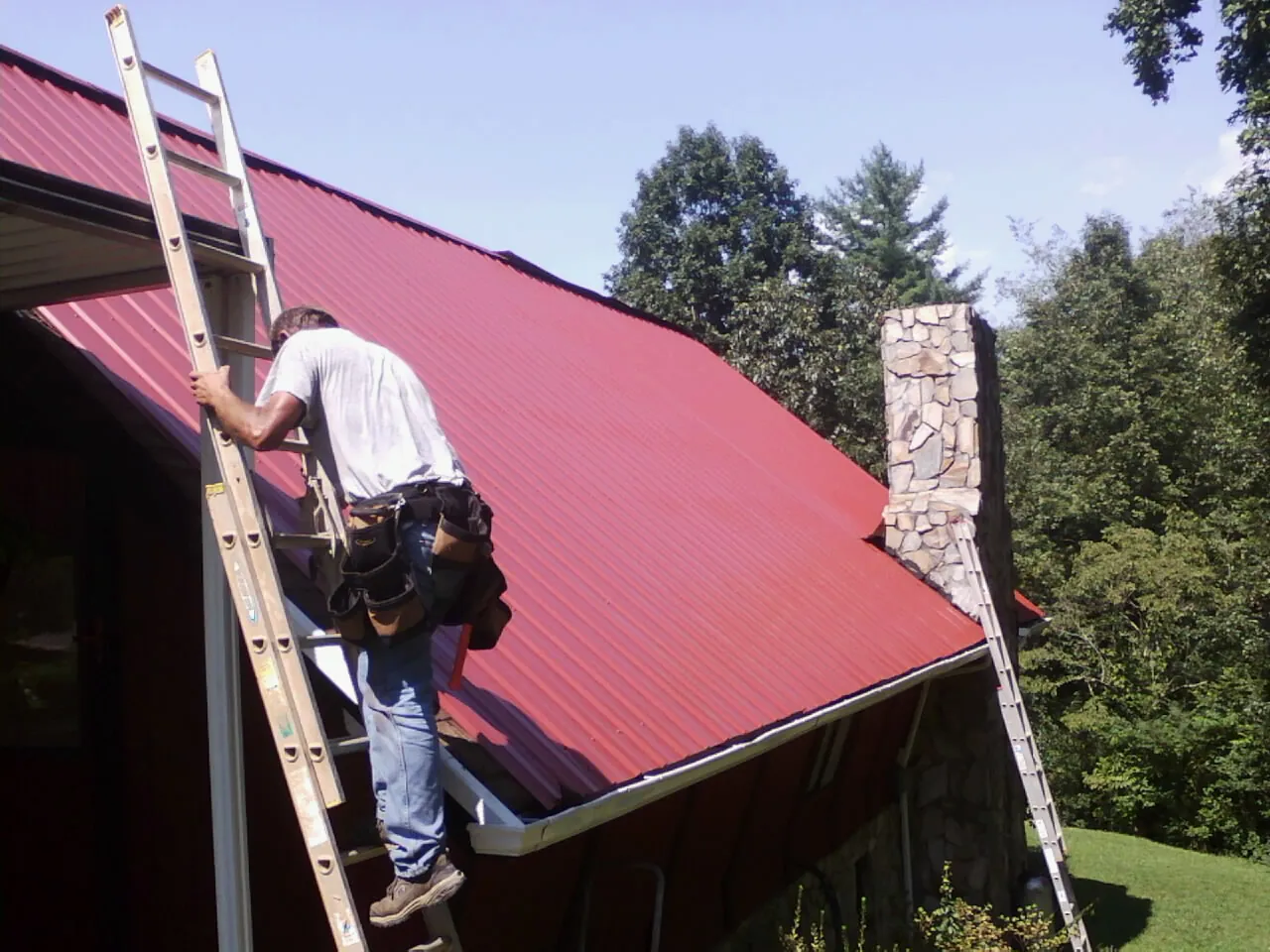 Roofer on a ladder beside a red metal roof and stone chimney — Soffit & Fascia Repair by Roofing Brothers in Sherburne
